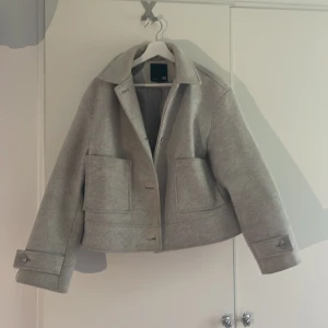 Grå kappa - Snygg grå jacka från lager 157 i ullblandning med  fickor framtill och krage. Jackan är croppad och har knappar både framtill och vid ärmsluten. Skicketr bra men finns tecken på användning. En söm i innertyget har gått upp, det går dock att sy igen. Ny: 400kr