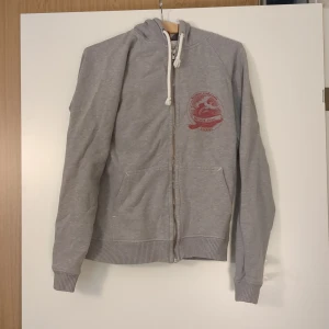 Grå hoodie med tryck och dragkedja Encore Storlek S - Grå hoodie med vit dragsko i huvan och röd logga på bröstet. Tröjan har hel dragkedja framtill, kängurufickor och ribbade muddar vid ärmslut och nederkant. Perfekt för chill dagar eller när du vill ha en avslappnad stil.