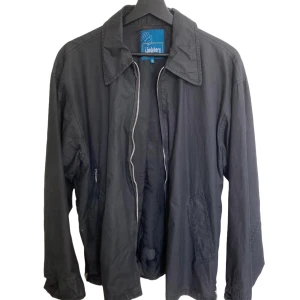 Svart vindjacka från J.Lindeberg - Svart vindjacka från J.Lindeberg med klassisk krage och dragkedja framtill. Jackan har två snedställda fickor och är tillverkad i ett lätt material som passar perfekt för blåsiga dagar. Enkel och stilren design med lång ärm. En defekt på insidan längst ner, synlig i bilden. Syns dock inte från utsidan. I övrigt är den i bra skick.