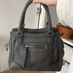 Grå handväska med snygga detaljer - Sparsamt använd väska som tyvärr inte kommer till användning längre💕Stilren grå handväska med två rejäla handtag och coola flätade detaljer på sidorna. Väskan har dragkedja framtill och baktill för extra förvaring. Pris kan diskuteras vid snabb affär!☺️