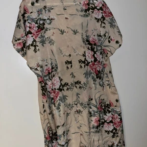 Beige blommig kortklänning i satin - Superfin beige kortklänning med blommigt mönster i rosa, grönt och vitt. Klänningen har rund halsringning, korta ärmar och är gjord i ett glansigt satinliknande material som känns lyxigt och flowy. Perfekt för dig som gillar romantiska vibes och mjuka färger.