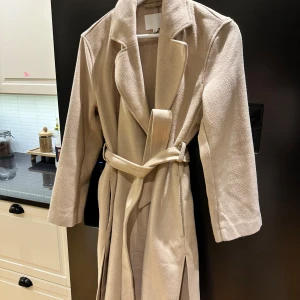 Beige kappa med bälte och krage - Stilren beige kappa med klassisk krage och bälte i midjan. Jackan har långa ärmar och en rak, lite loose passform. Materialet är mjukt och känns som ullblandning. Perfekt för dig som gillar en clean och tidlös look.