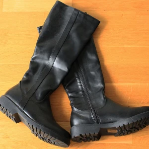 Svarta stövlar - Svarta knähöga stövlar med grov sula och dragkedja på insidan. Skorna är tillverkade i skinnimitation och har en rund tå. Perfekta för en stilren look till höst och vinter. Har aldrig blivit använda.