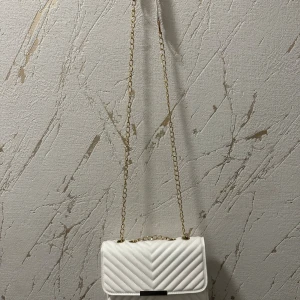 Vit quiltad axelväska med kedja - Snygg vit axelväska med quiltat mönster och guldfärgad kedjerem. Väskan har ett stilrent, rektangulärt format och stängs med magnetlås. Perfekt för dig som vill ha en trendig och elegant accessoar till din outfit.