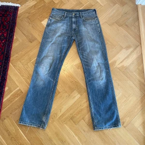 Blå raka jeans från Carhartt - Säljer ett par klassiska blå jeans från Carhartt med raka ben och slitna detaljer. Jeansen har fem fickor, bälteshällor och en snygg läderpatch på bakfickan. Perfekt för dig som gillar en avslappnad och tidlös look. Sanna storleken är 32/33
