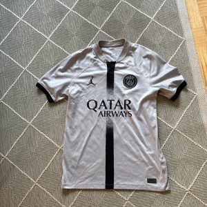 PSG x Jordan fotbollströja grå M - Snygg PSG fotbollströja i grått med svart vertikal rand och svarta detaljer. Jordan-logga på bröstet och Paris Saint-Germain-emblem. Tillverkad i lätt och ventilerande material, perfekt för träning eller match. Qatar Airways tryck på framsidan.