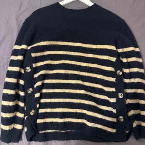 Randig stickad tröja med knappar Indiska  - Mysig stickad tröja i mörkblått och beige med breda ränder. Tröjan har lång ärm och coola knappar längs sidorna som extra detalj. Perfekt för lager-på-lager och chill stil.