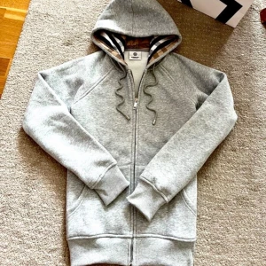 Grå hoodie från Burberry med dragkedja - Snygg grå hoodie från Burberry med klassiskt rutigt foder i huvan och broderad logga på bröstet. Tröjan har hel dragkedja, snörning i huvan och är tillverkad i 100% bomull. Perfekt för en chill och stilren look.