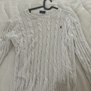 Vit kabelstickad tröja Polo Ralph Lauren - Vit kabelstickad tröja från Polo Ralph Lauren med klassisk rund hals och broderad marinblå logga på bröstet. Tröjan är långärmad och har ribbade muddar vid ärmslut och nederkant. Perfekt för lager-på-lager under hösten och vintern. Storlek L men sitter som M