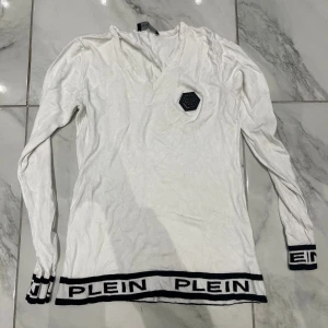 Vit v-ringad tröja  - Snygg vit långärmad tröja v-ringning och svart logga på bröstet. Tröjan har svarta detaljer. Mjuk och stretchig känsla, perfekt för en trendig look.