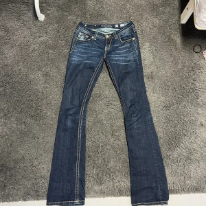 Miss Me bootcut jeans med broderi - Mörkblå bootcut jeans från Miss Me med snygga slitningar och broderade detaljer på bakfickorna. Tydliga kontrastsömmar och dekorativa knappar ger jeansen en unik look. Perfekt för dig som gillar jeans med attityd och detaljer. Strl 28 med längden 36 så passar tjejer med långa ben