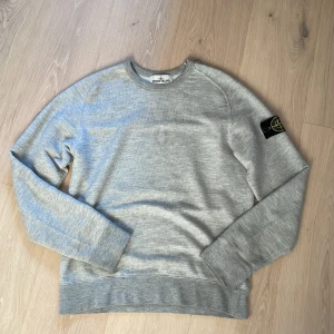 Grå sweatshirt från Stone Island - Säljer en stilren grå sweatshirt från Stone Island med klassisk rund hals och ikonisk patch på vänster ärm. Tröjan är i mjuk bomull och har ribbade muddar vid ärmslut och nederkant. Perfekt för dig som gillar streetwear och enkel stil.