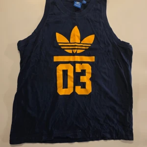 Mörkblå Adidas linne med gul logga - Säljer ett mörkblått linne från Adidas. Linnet är ärmlöst och har en sportig look, perfekt för dig som gillar streetwear. Tillverkat i mjuk bomull för skön och snygga känsla.