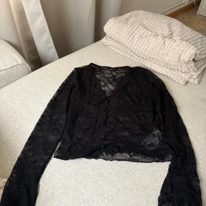 Svart spetsblus från Gina Tricot - Säljer en svart blus från Gina Tricot i storlek M. Blusen är helt i spets med blommigt mönster, v-ringad och har långa ärmar. Den har knappar framtill och är lätt transparent, vilket ger en snygg och trendig look.