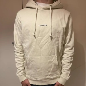 Vit hoodie från Les Deux med tryck - Säljer en stilren vit hoodie från Les Deux med svart broderad logga på bröstet. Tröjan har huva med snörning, magficka och ribbade muddar. Perfekt för dig som gillar enkel och clean streetstyle.