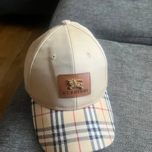 Beige Burberry keps med rutigt mönster - Snygg beige keps från Burberry med klassiskt rutigt mönster på skärmen och läderpatch med guldlogga framtill. Justerbar metallspänne bak med Burberry-gravyr. Kepsen är i bomull och har broderad Burberry-text på sidan.
