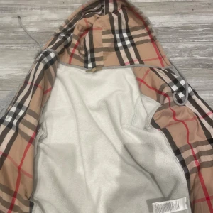 Grå hoodiejacka från Burberry - Grå hoodiejacka från Burberry med klassiskt rutigt foder i beige, svart, vitt och rött. Jackan har dragkedja framtill, huva med snörning och broderad logga på bröstet. Tillverkad i mjuk bomull med elastan för extra komfort.