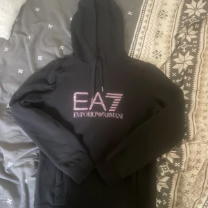 Svart EA7 Emporio Armani hoodie - Svart hoodie från EA7 Emporio Armani med stor logga i glansig lila på bröstet och ryggen. Klassisk huva med snörning och långa ärmar. Snygg😀