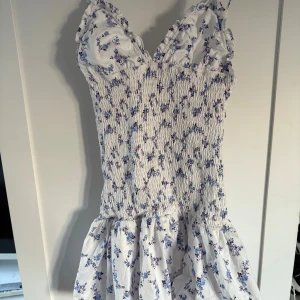 Vit blommig kortklänning med volang - Supersöt vit kortklänning med blått blommönster och smockad överdel. Klänningen har volangiga axelband och en luftig kjol med volangkant. Perfekt för varma dagar och ger en romantisk vibe.