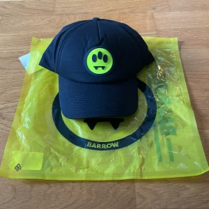 Svart keps från Barrow med smiley - Svart keps från Barrow med justerbar rem bak och broderad logga. Framtill finns Barrows ikoniska neongula smiley med vampyrtänder. Kepsen är i bomull och har en klassisk böjd skärm. Perfekt för dig som vill sticka ut med en cool detalj. Ny pris 1500kr och den finns inte kvar på deras hemsida, pris kan diskuteras 