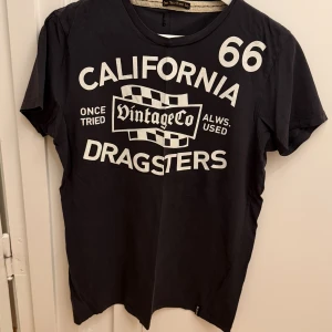 Mörkgrå t-shirt med vitt tryck - Mörkgrå t-shirt med stort vitt tryck på framsidan: 'California 66 Dragsters' och schackrutigt motiv. Klassisk rund hals och korta ärmar. Skön och mjuk bomullskvalitet, perfekt för en avslappnad streetstil. Storlek M.