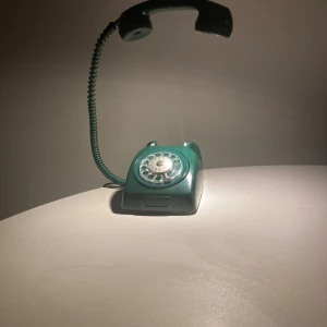 Grön retro bordstelefon med nummerskiva - Klassisk grön bordstelefon med roterande nummerskiva och spiralsladd. Telefonen har en robust design och tydliga knappar. Mindre repor och bruksspår syns, men överlag gott skick. Perfekt för retroälskare eller som inredningsdetalj.