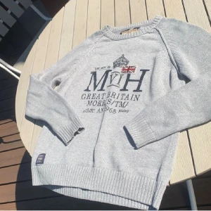 Grå stickad tröja från Morris - Snygg grå stickad tröja från Morris med tryck och brittisk flagga framtill. Tröjan har rund halsringning, långa ärmar och ribbade muddar. Perfekt för dig som gillar klassisk stil med en sportig touch.