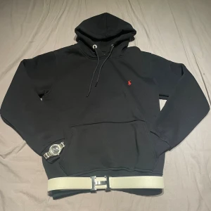 Svart Ralph Lauren hoodie - Svart hoodie från Ralph Lauren med klassisk röd broderad logga på bröstet. Tröjan har huva med snörning, känguruficka och ribbade muddar. Perfekt för dig som gillar en clean och stilren look.