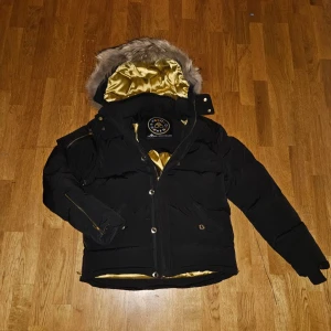 Moose Knuckles pufferjacka - Säljer en svart pufferjacka från Moose Knuckles med guldfärgat, glansigt foder och stor huva med fluffig päls. Jackan har dragkedja, knappar och flera fickor. Perfekt för kalla dagar och riktigt snygg detalj med guldfodret. Pris kan diskuteras vid intresse.