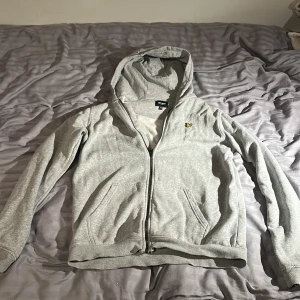 Grå hoodie från Lyle & Scott Junior - Grå hoodie från Lyle & Scott Junior med dragkedja och huva. Klassisk gul logga på bröstet och två fickor framtill. Mjuk bomullskänsla och normal passform, perfekt för chill dagar eller när du vill ha en enkel och stilren look.