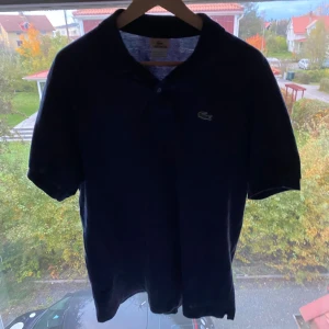 Mörkblå pikétröja från Lacoste - Snygg mörkblå pikétröja från Lacoste med klassisk krage och korta ärmar. Tröjan har den ikoniska gröna krokodilloggan på bröstet och är tillverkad i 100% bomull för en skön och luftig känsla. Trasig knapp som man ser på sista bilden