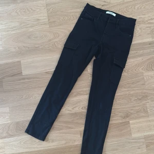 Svarta jeans med fickorna på sidan från Kappahl strl 36/S - Svarta cargopants från Kappahl med raka ben och klassiska cargofickor på sidorna. Byxorna har bälteshällor, femficksdesign och är tillverkade i ett mjukt bomullsmaterial med lite stretch. Perfekta för en avslappnad och trendig look.