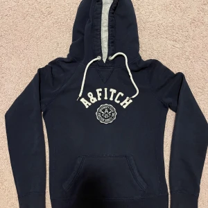Mörkblå hoodie Abercrombie & Fitch S - Mörkblå hoodie från Abercrombie & Fitch med broderad logga och text på bröstet. Tröjan har vit snörning i huvan, känguruficka och mjukt grått foder i huvan. Perfekt för chill dagar och snygg streetstil.