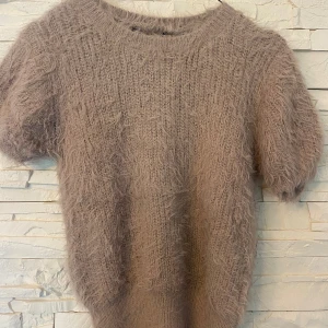 Beige fluffig stickad tröja - Supermjuk och fluffig stickad tröja i beige med korta ärmar och rund hals. Perfekt för dig som gillar en cozy och trendig look. Tröjan har en lös passform och är tillverkad i ett lurvigt material som känns extra mysigt.