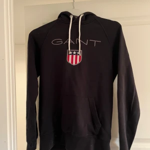 Svart hoodie från GANT med logga XS - Svart hoodie från GANT i storlek XS med vit snörning i huvan och broderad GANT-logga samt sköld på bröstet. Klassisk känguruficka framtill och långa ärmar. Perfekt för dig som gillar stilren och sportig look.