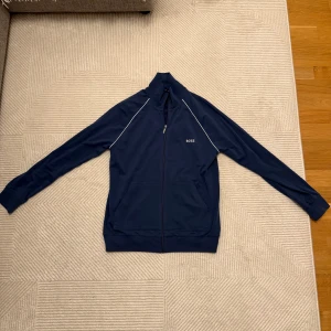 Marinblå ziptröja från BOSS - Snygg marinblå ziptröja från BOSS med vita detaljer längs axlarna och diskret logga på bröstet. Tröjan har hel dragkedja, hög krage och två fickor framtill. Perfekt för en sportig och clean look.