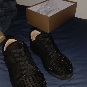 Christian Louboutin svarta sneakers med nitar - Svarta sneakers från Christian Louboutin i mocka med svarta nitar på tån och klassisk röd sula. Skorna har snörning och rund tå, samt exklusiva detaljer som ger en edgy vibe. Perfekta för dig som vill sticka ut med lyxig streetstyle.