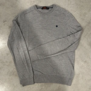 Morris tröja  - Snygg grå stickad tröja från Morris med rund halsringning och diskret broderad logga på bröstet. Tröjan är i mjuk merinoull och har ribbade muddar vid ärmslut och nederkant. Perfekt för lager-på-lager under hösten och vintern. PASSAR M
