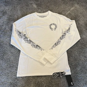Vit långärmad Chrome Hearts tröja - Vit långärmad tröja från Chrome Hearts med svart tryck på bröstet och längs ärmarna. Snyggt grafiskt mönster och rund hals. Materialet är mjuk bomull och tröjan har en clean och modern vibe. Perfekt för dig som gillar streetwear och exklusiva märken.