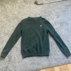 Mörkgrön stickad tröja från Morris - Snygg mörkgrön stickad tröja från Morris i storlek M. Tröjan har rund halsringning, ribbade muddar och ett broderat Morris-märke på bröstet. Perfekt för kyliga dagar och enkel att matcha med jeans.