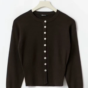 Brun kofta från Gina Tricot - Mörkbrun, ribbstickad kofta från Gina Tricot med långa ärmar och vita knappar framtill. Modellen är croppad och har en tight passform som framhäver midjan. Perfekt för lager-på-lager och snygg till både jeans och kjol. och helt oanvänd 