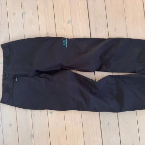 Svarta Gore-Tex byxor State of Elevenate XS - Svarta skalbyxor från State of Elevenate i storlek XS. Byxorna har diskret logga i turkos, justerbar midja med kardborre och dragkedjor vid bensluten. Perfekta för som skalbyxor höst & vår med mellanlager under eller som regnbyxor under sommaren och designade i Åre, Sverige.