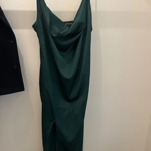 Mörkgrön långklänning från Nelly - Snygg mörkgrön långklänning med draperad urringning och slits framtill. Klänningen är ärmlös och har en elegant, slät yta som ger en lyxig känsla. Perfekt för dig som vill sticka ut med en stilren look.