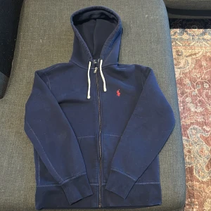 Marinblå hoodie från Polo Ralph Lauren - Snygg marinblå hoodie från Polo Ralph Lauren med dragkedja och vit snörning i huvan. Klassisk röd broderad logga på bröstet. Mjuk bomullskvalitet och känguruficka framtill. Perfekt för chill dagar eller när du vill ha en clean streetstyle.