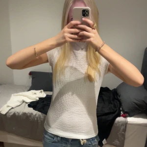 Vit strukturerad topp med kort ärm - Säljer en vit topp med korta ärmar och snyggt strukturerat tyg. Modellen är figurnära och har en enkel, stilren look som passar till jeans eller kjol. Tyget är lätt och känns skönt mot huden. Perfekt för dig som gillar en clean och fräsch stil.