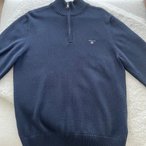 Marinblå tröja från Gant - Snygg marinblå stickad tröja från Gant med halv dragkedja och ribbad krage. Klassisk logga broderad på bröstet. Perfekt för lager-på-lager och passar till både jeans och chinos.