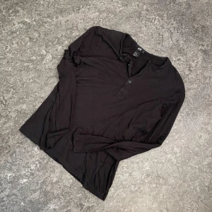 Svart långärmad pikétröja H&M - Svart långärmad pikétröja från H&M i mjuk bomull. Tröjan har knappar vid halsen och en enkel, clean look som passar till allt. Perfekt för dig som gillar en basic stil med lite edge.