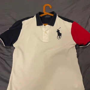 Pikétröja Polo Ralph Lauren, vit/blå/röd - Snygg pikétröja från Polo Ralph Lauren i vit, marinblå och röd. Stor broderad logga framtill, stor siffra 3 på ryggen och amerikansk flagga på ärmen. Klassisk krage och korta ärmar. 