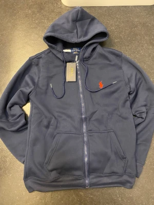 Polo Ralph Lauren blå zip hoodie - Stilren blå zip hoodie från Polo Ralph Lauren med klassisk logga på bröstet. Mjuk och bekväm passform, perfekt till vardags. Storlek-S 