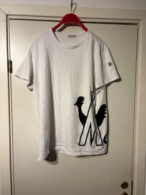 Vit Moncler t-shirt med tryck - Säljer en clean vit t-shirt från Moncler med svart grafiskt tryck framtill och stor logga baktill. Klassisk rund hals, korta ärmar och ikonisk Moncler-patch på ärmen. Tillverkad i mjuk bomull som känns skön mot huden.